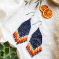 A pair of long beaded dangle earrings with a navy blue top section and a tapered fringe in orange, yellow, and white, displayed on a textured white fabric with dried orange slices, a succulent, and small decorative beads.