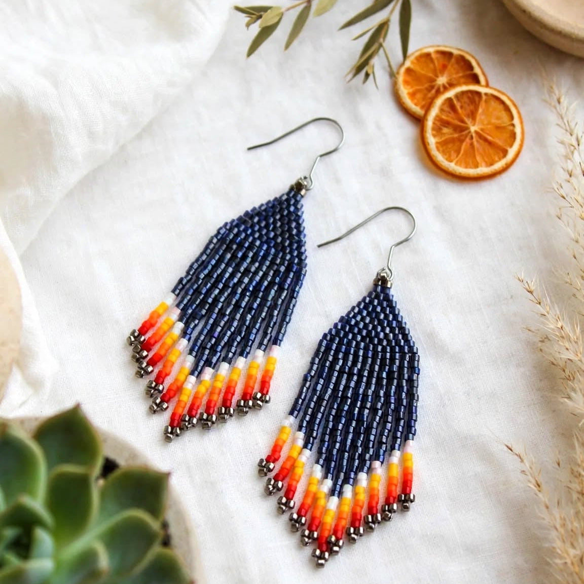 A pair of long beaded dangle earrings with a navy blue top section and a tapered fringe in orange, yellow, and white, displayed on a textured white fabric with dried orange slices, a succulent, and small decorative beads.