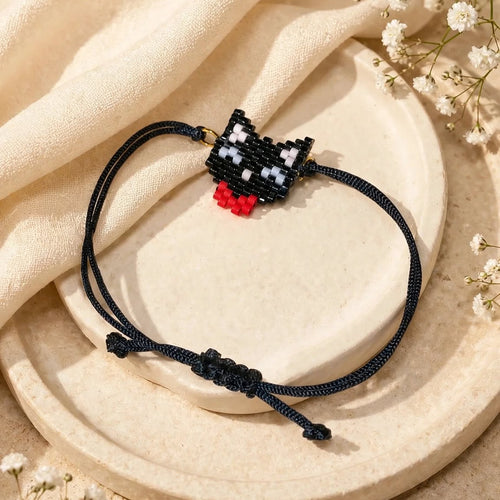 A black cord bracelet with a small black-and-white beaded cat charm and a red accent bead, displayed on a beige ceramic plate with draped cream fabric and small white flowers.