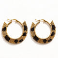 A pair of gold-tone hoop earrings with a textured, scalloped edge featuring alternating rows of small gold and black beads, displayed on a clean white background.