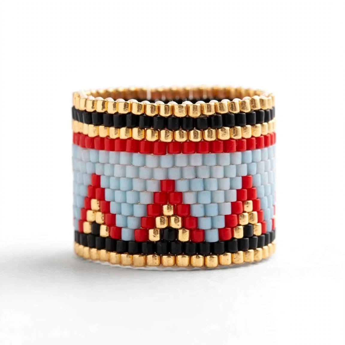 A wide beaded ring featuring a geometric pattern with red, black, and light blue triangles on a white base, bordered by gold beads, set against a plain white background.