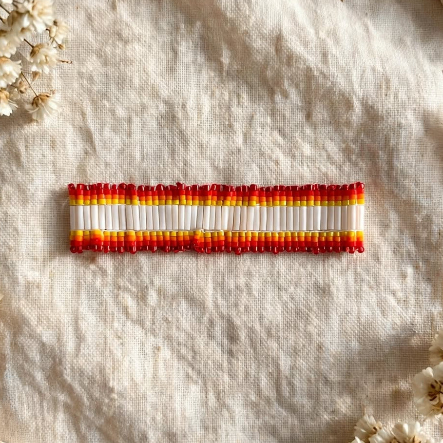 A small rectangular woven or beaded accessory with horizontal stripes in red, orange, yellow, and white, placed on a textured cream fabric with dried flowers and stone props around it.