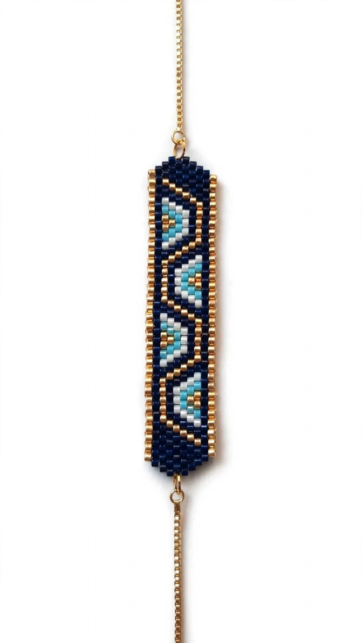 A beaded geometric stretch bracelet featuring a navy blue base with light blue and gold patterned accents, suspended from a gold chain on a white background.