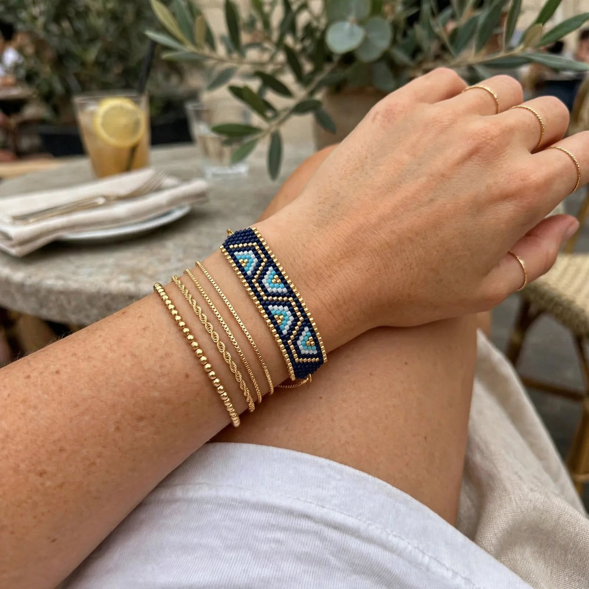 A close-up of a person's wrist wearing a beaded geometric bracelet with a navy blue and turquoise pattern, layered with several gold-tone chain bracelets, in an outdoor cafe setting.