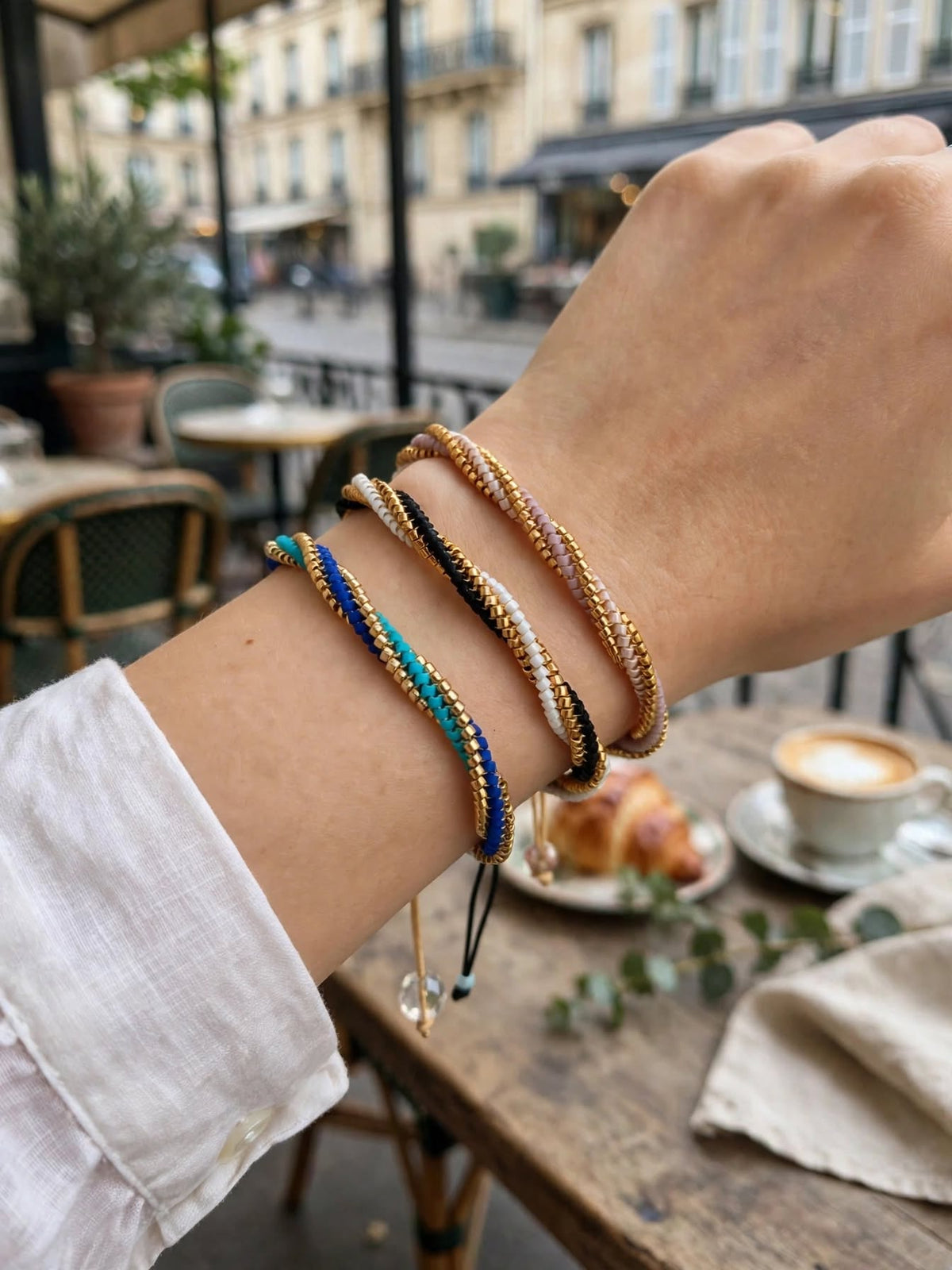 A close-up of a person's wrist wearing four braided cord bracelets in different colors, set against a blurred background of a cafe table with a coffee cup and croissants.