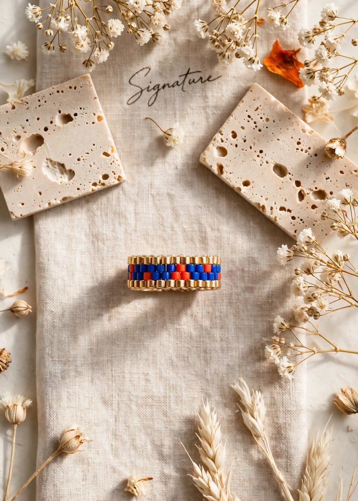 A handmade beaded bracelet with blue, red, and gold beads is displayed on a beige linen cloth, surrounded by dried flowers, pampas grass, and two decorative stone tiles. The brand name 'Signature' is written in cursive in the background.