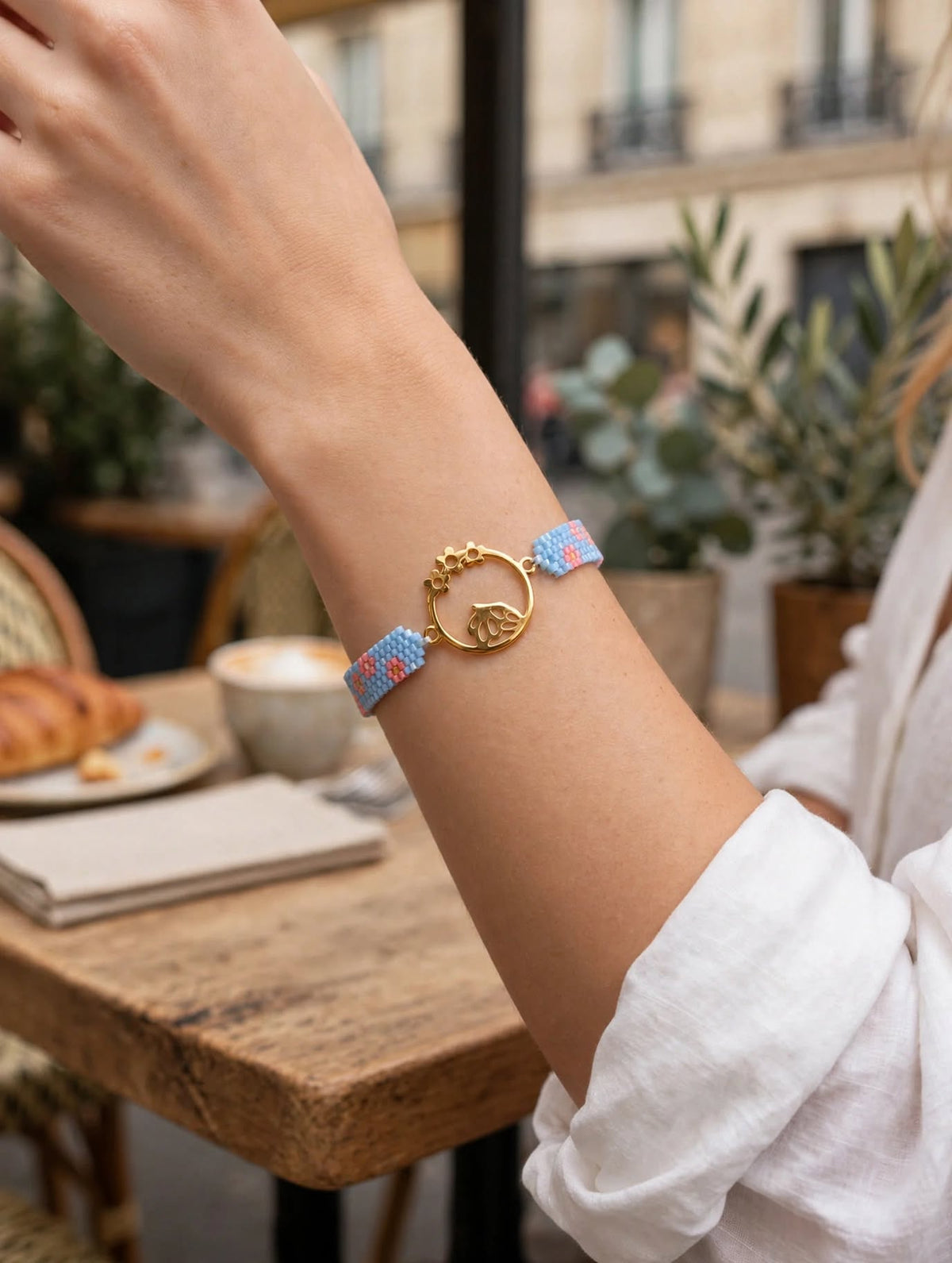 A gold-plated beaded bracelet with a central circular charm and a small gold heart, worn on a person's wrist in an outdoor cafe setting.