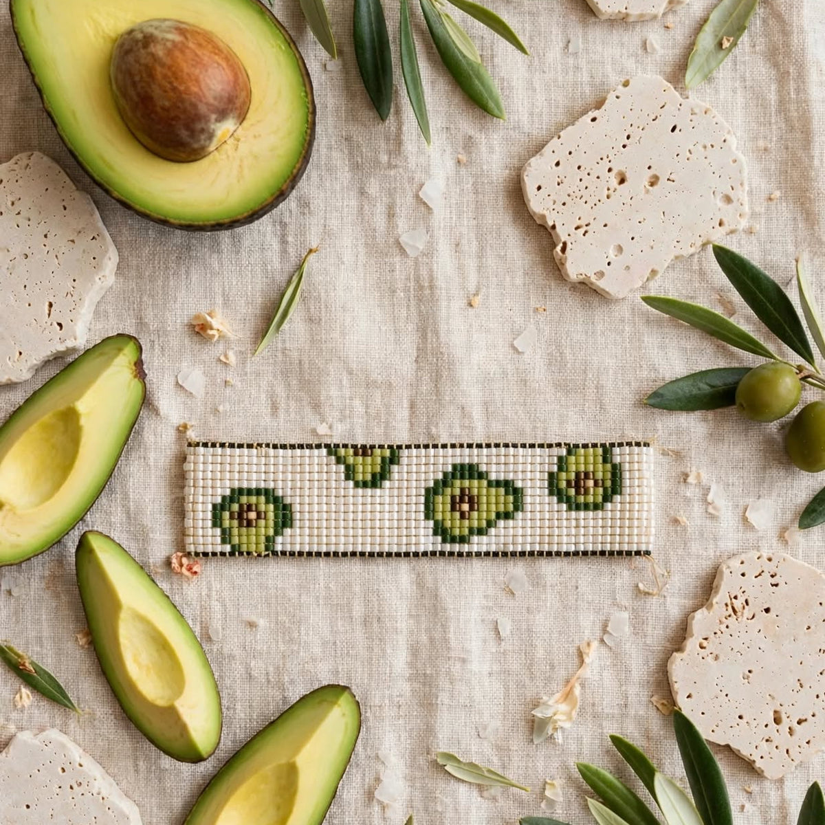 A rectangular beaded bracelet with a green avocado pattern on a cream background, styled in a flat-lay with fresh avocado halves, olive branches, and stone crackers on a linen surface.