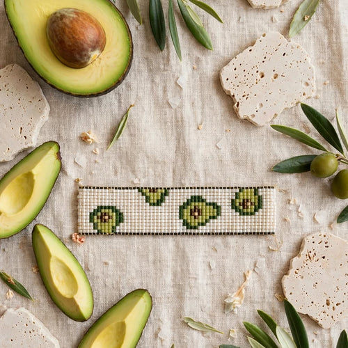A rectangular beaded bracelet with a green avocado pattern on a cream background, styled in a flat-lay with fresh avocado halves, olive branches, and stone crackers on a linen surface.