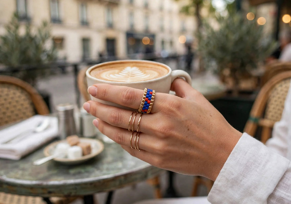 A close-up lifestyle shot of a person's hand holding a cup of latte at an outdoor cafe, wearing a gold ring with blue and red beads on one finger and two slim gold bands on the other hand.