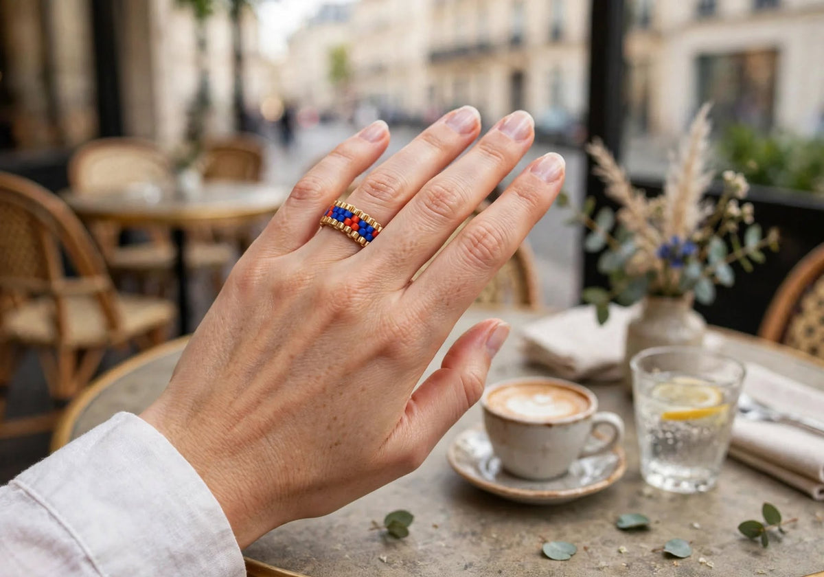 A close-up of a hand wearing a gold ring with blue and red geometric accents, set in a cozy cafe environment with a coffee cup, glass of water, and blurred background tables.