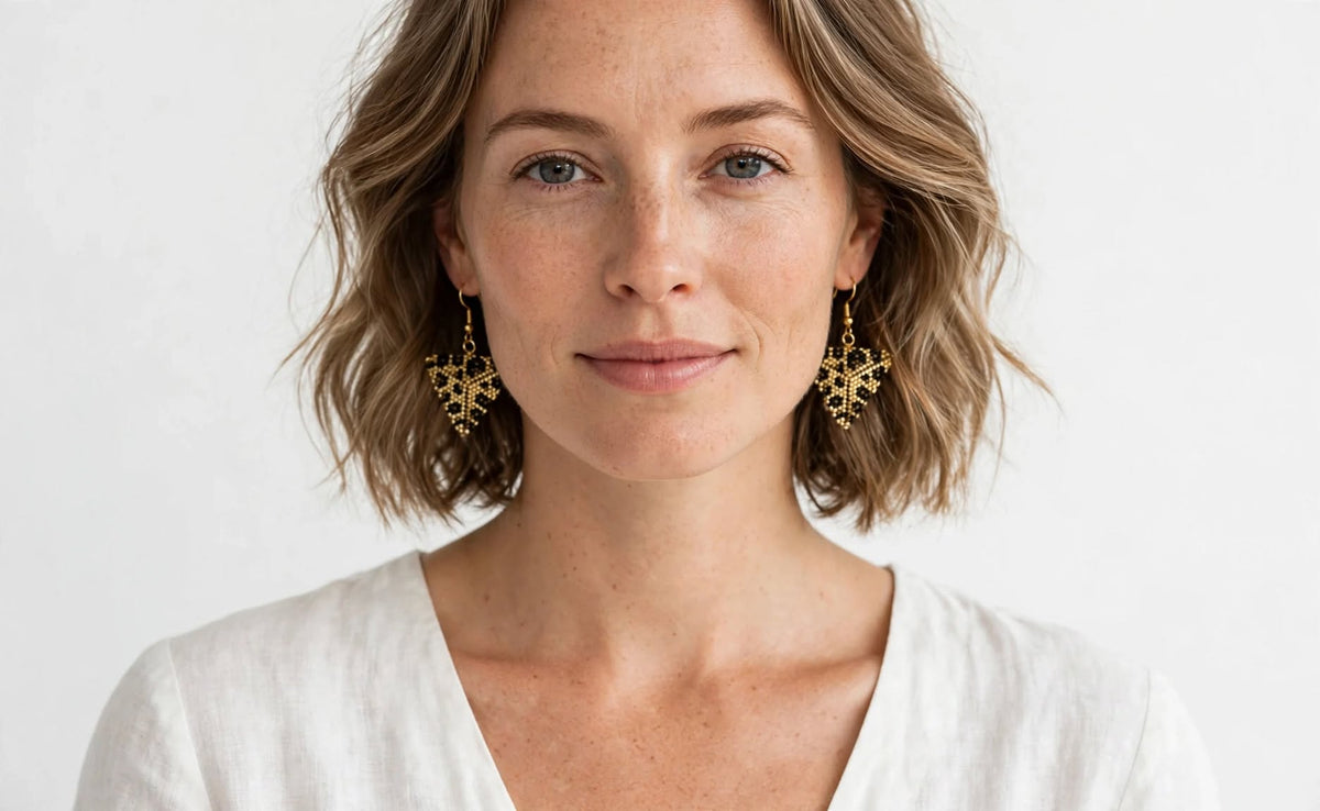 A woman with short wavy light brown hair wearing ornate gold dangle earrings and a white V-neck top, photographed against a clean white background.