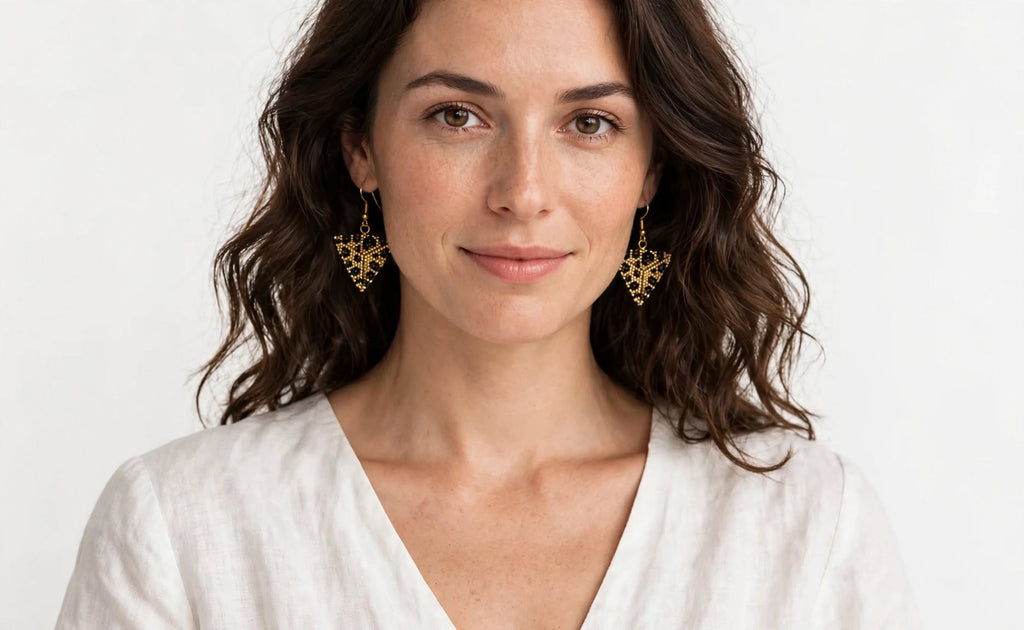 A woman with wavy brown hair wearing gold dangling earrings and a white V-neck top, photographed against a clean white background.