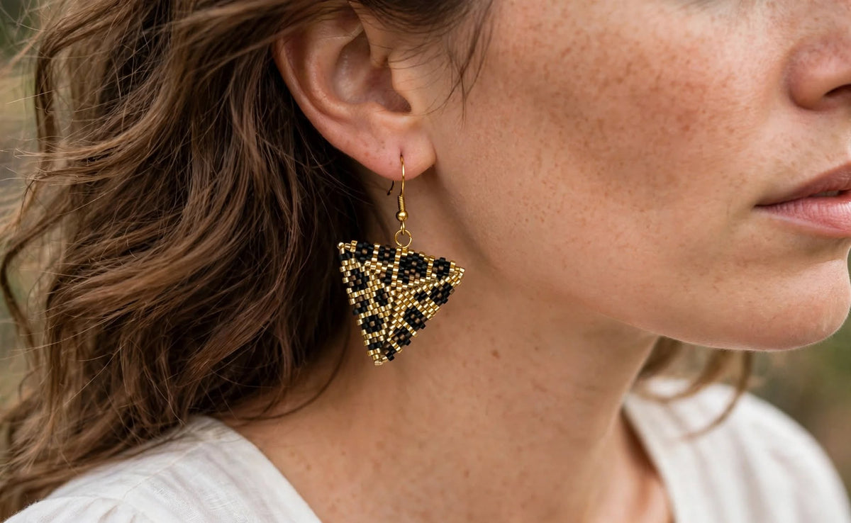 A close-up of a woman wearing a triangular beaded dangle earring in black and gold, with her ear, hair, and part of her face visible.