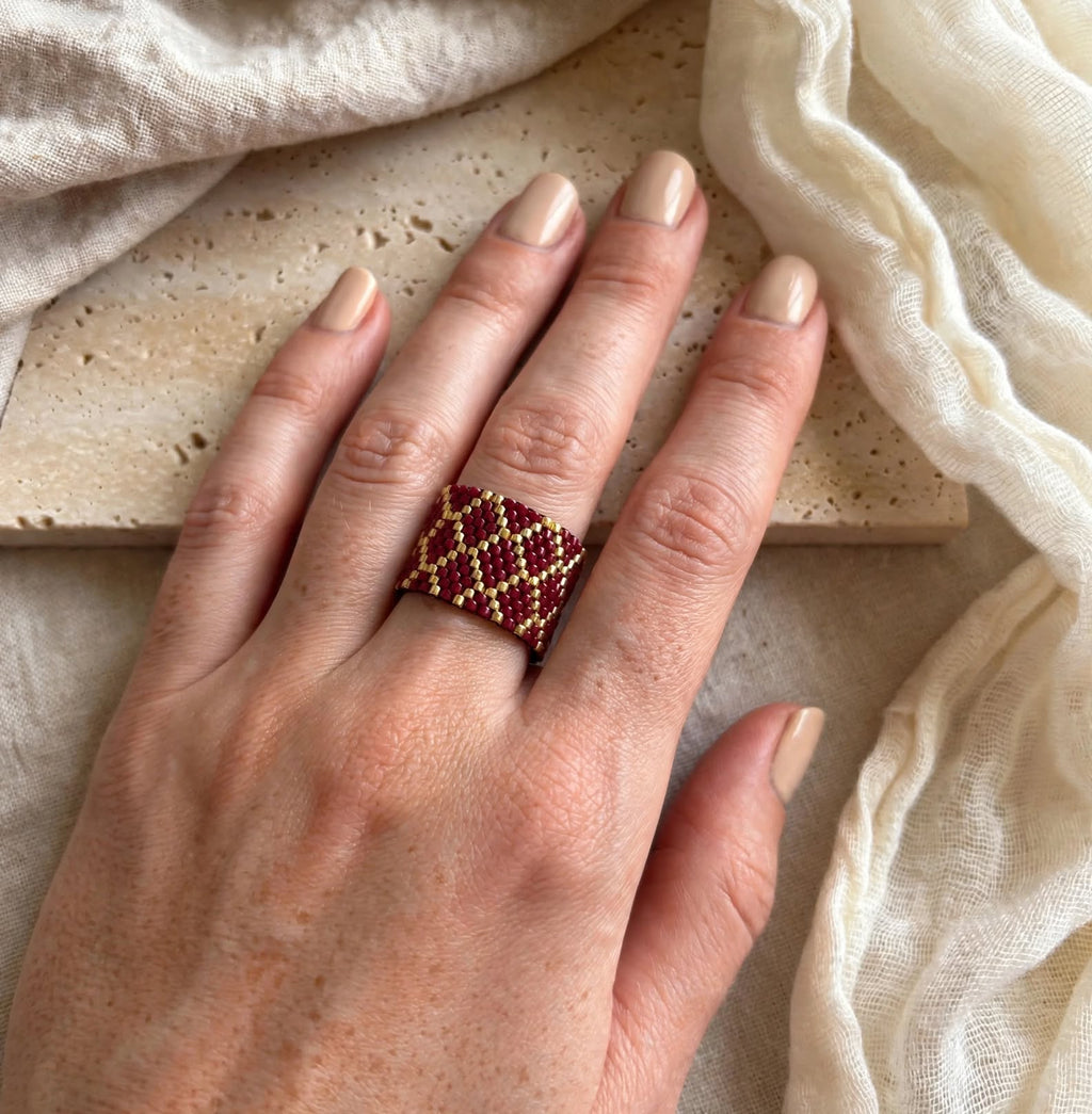 A close-up of a hand wearing a woven beaded ring with a geometric diamond pattern in deep red and gold, styled with neutral beige fabric and a stone slab background.
