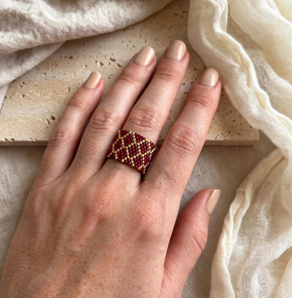 A close-up of a hand wearing a woven beaded ring with a geometric diamond pattern in deep red and gold, styled with neutral beige fabric and a stone slab background.