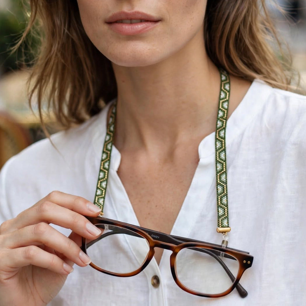 A woman in a white blouse holds a pair of tortoiseshell eyeglasses with a green patterned neck strap against a softly blurred outdoor cafe background.