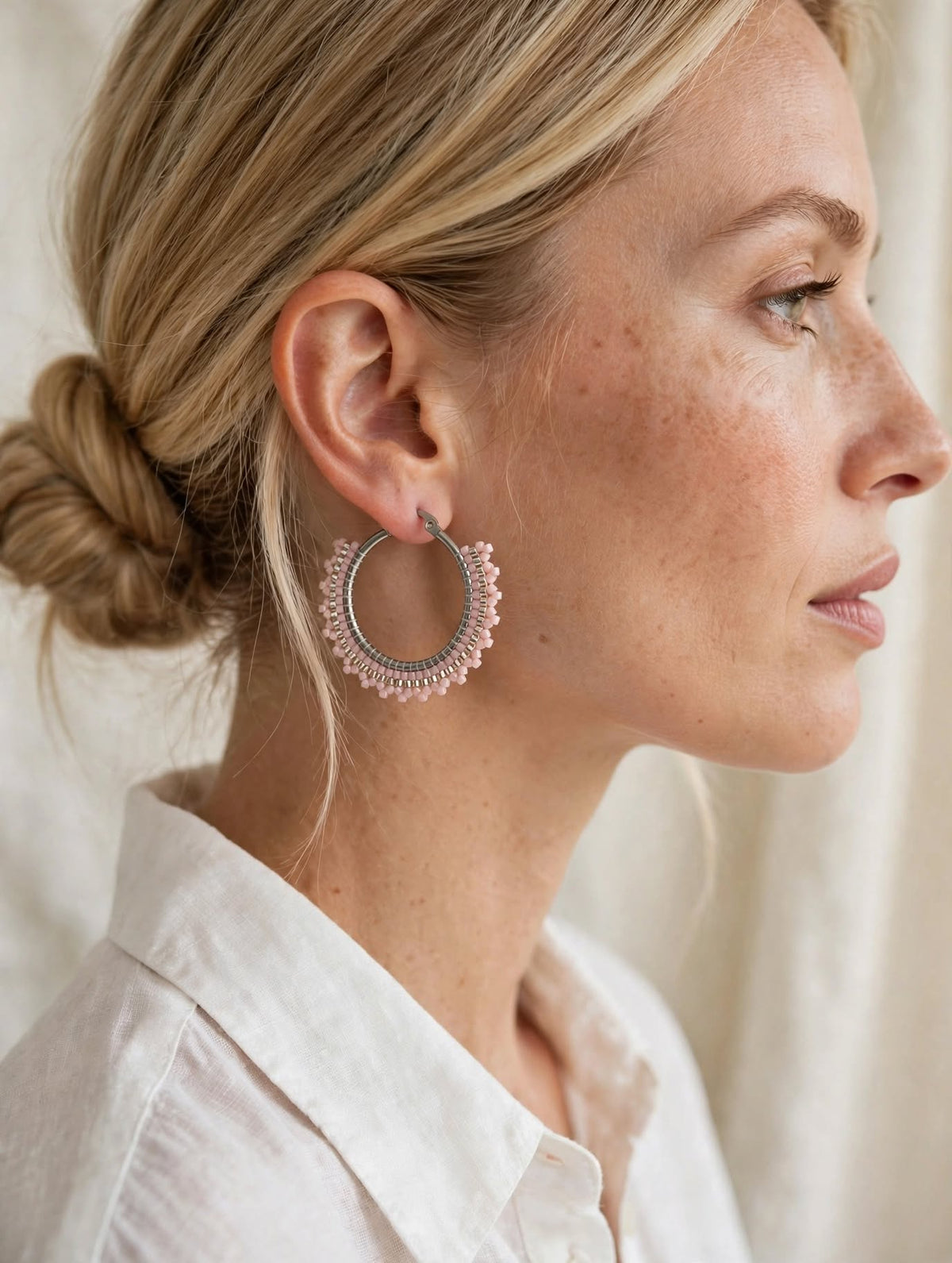 A close-up side profile of a woman with blonde hair in a low bun, wearing a large circular silver hoop earring adorned with delicate pink floral lace detailing, paired with a white collared shirt.