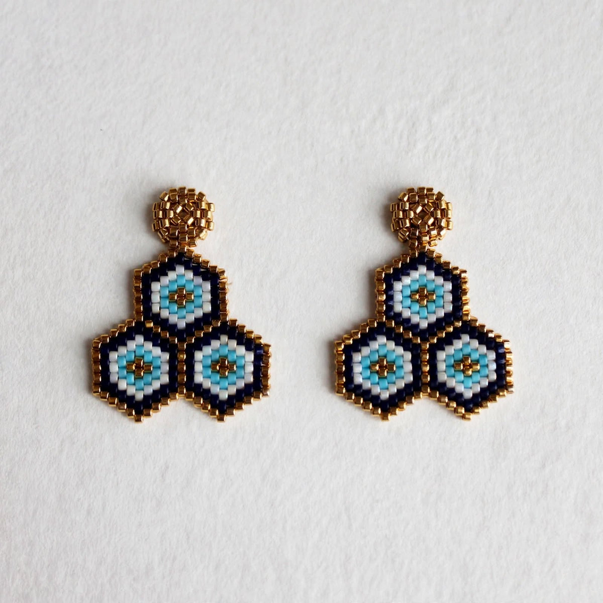 A pair of handmade beaded geometric dangle earrings featuring three nested hexagonal motifs in light blue, white, and black, finished with textured gold-tone studs, displayed on a clean white background.