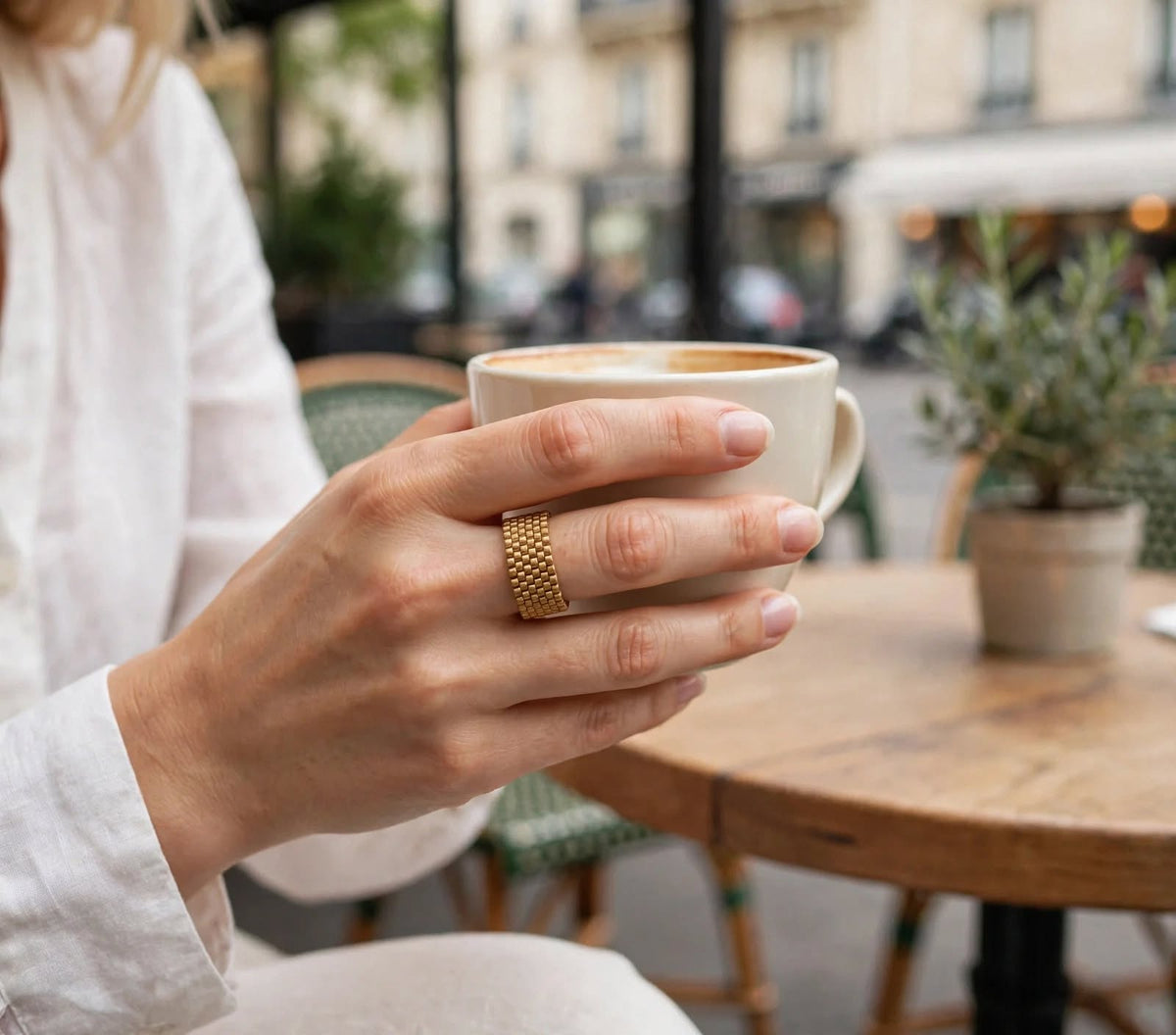 A close-up lifestyle shot of a person holding a cup of coffee at an outdoor cafe, wearing a textured gold band ring on their finger.