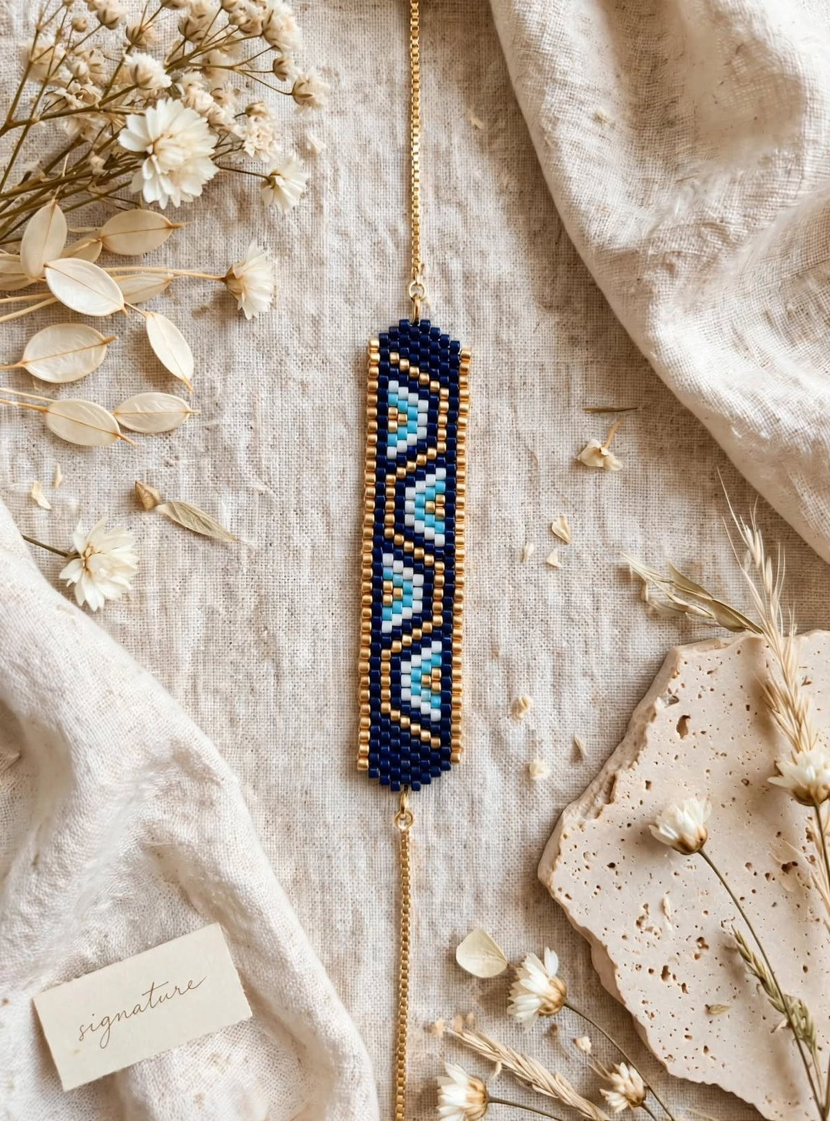 A bohemian-style beaded bracelet with a geometric pattern in navy blue, light blue, and white, suspended from a gold chain. The bracelet is displayed in a styled flat-lay arrangement with dried flowers, linen fabric, and ceramic tiles.