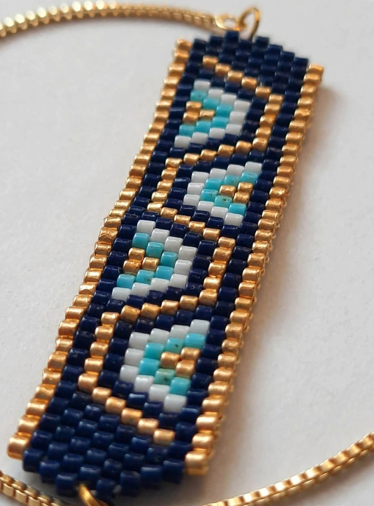 A close-up of a beaded geometric pendant necklace featuring navy blue, gold, light blue, and white seed beads, shown with a gold chain on a light gray background.