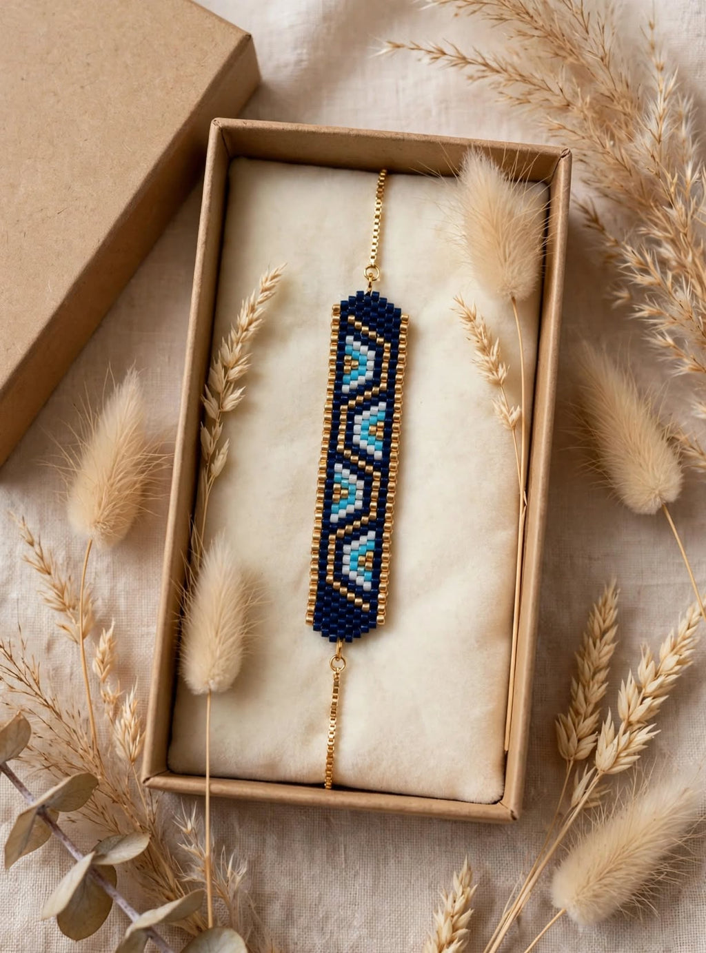 A beaded geometric charm bracelet with a navy blue, light blue, and gold pattern, presented inside a kraft gift box with dried wheat and bunny tail grass props.