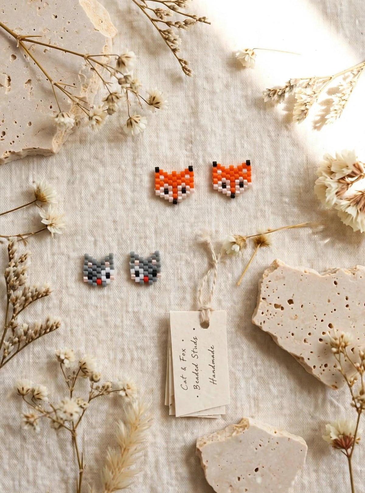 Handmade beaded stud earrings displayed on a textured cream fabric with dried flowers and stone chips. The set includes a pair of orange fox head earrings and a pair of gray cat head earrings, with a paper tag reading 'Cute & Fox - Baked Studs Handmade'.
