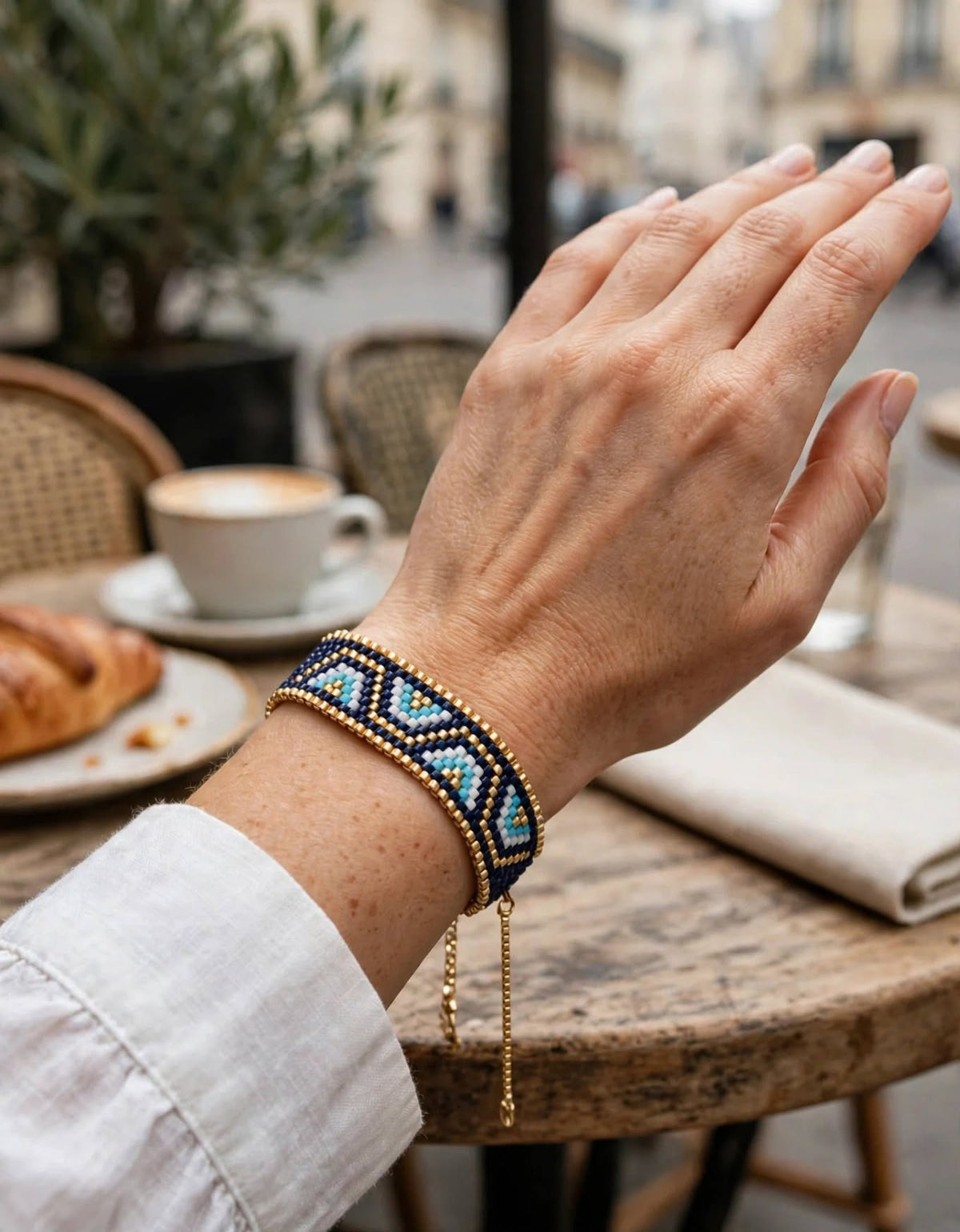 A close-up lifestyle shot of a person's hand wearing a beaded geometric bracelet with blue, white, and black beads and gold-toned chain drops, set against a blurred cafe background.