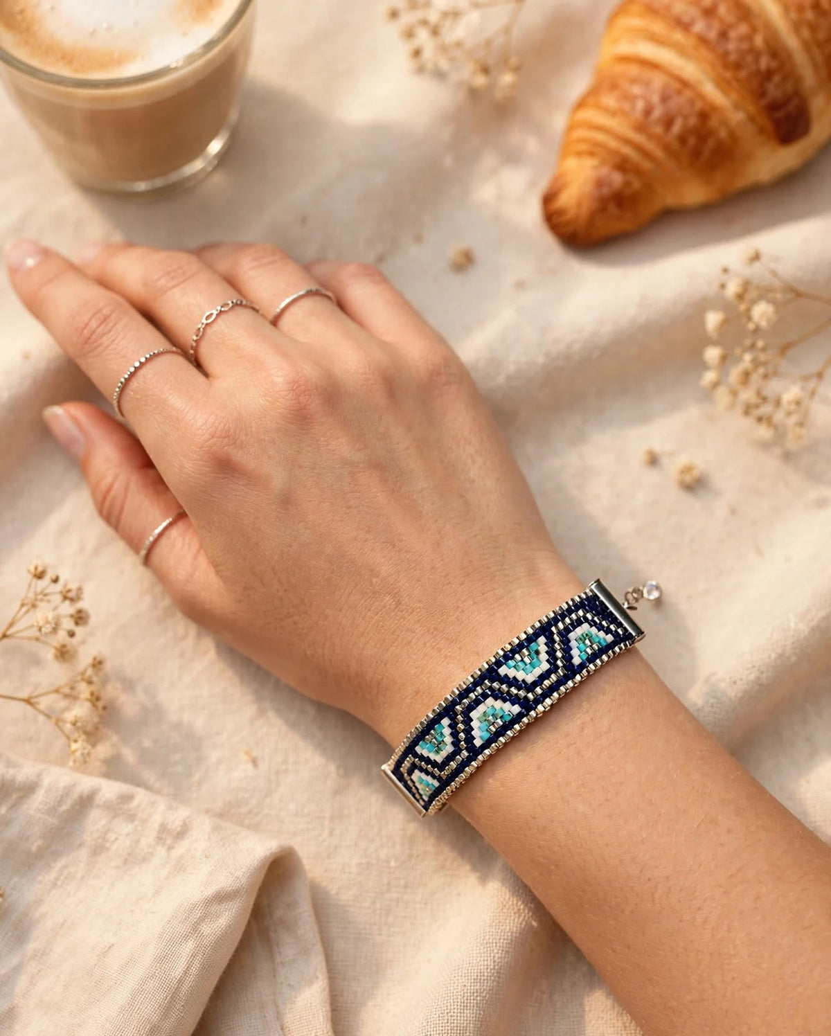A silver-toned beaded bracelet with a geometric pattern in turquoise and light blue, worn on a woman's wrist. The hand is resting on a beige fabric surface with a coffee cup, a croissant, and small dried flowers in the background.