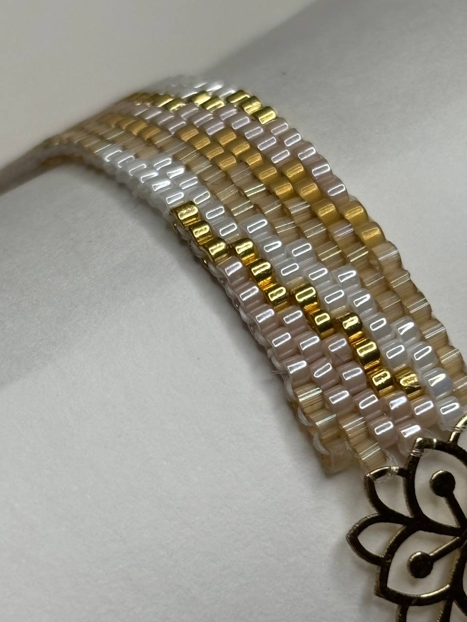 A close-up of a beaded bracelet displayed on a white mannequin wrist form. The bracelet features rows of small rectangular beads in white, clear, and gold, with a decorative gold-toned metal clasp visible at the bottom right.
