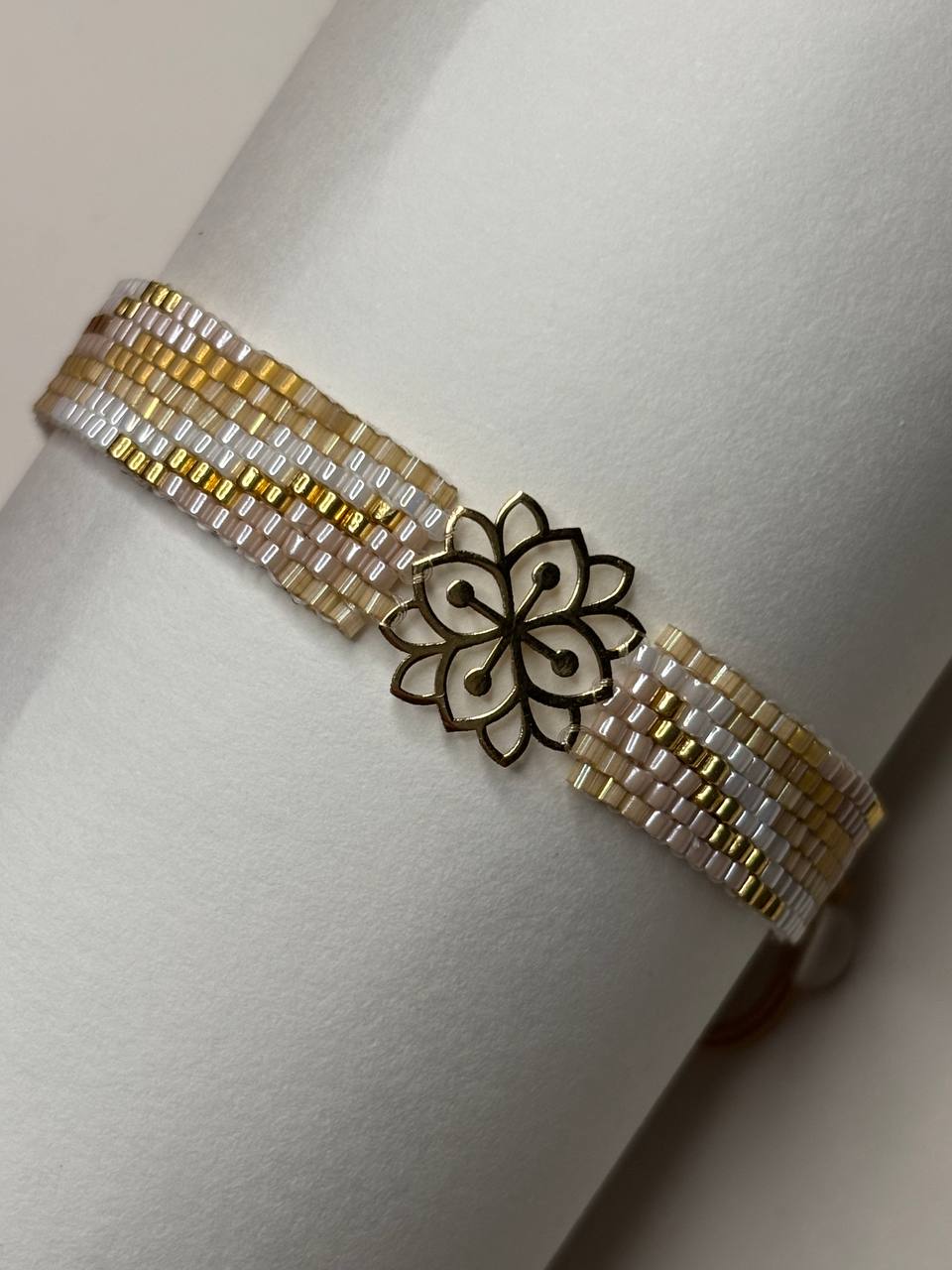 A beaded bracelet with white, gold, and silver beads, featuring a central black metal lotus flower charm, displayed on a white cylindrical stand.