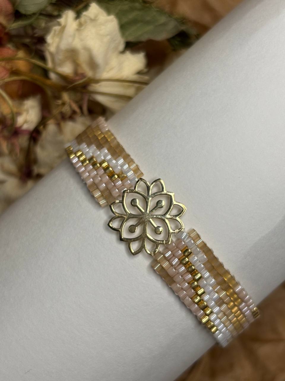 A beaded bracelet with a central gold-tone lotus flower charm, displayed on a white cylindrical stand. The bracelet features rows of small translucent beads in white, pale pink, and gold.