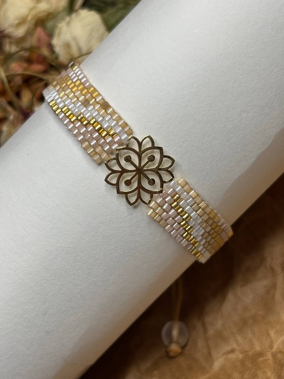 A beaded bracelet with white, gold, and beige seed beads, featuring a central gold-toned metal flower charm, displayed on a white cylindrical stand.