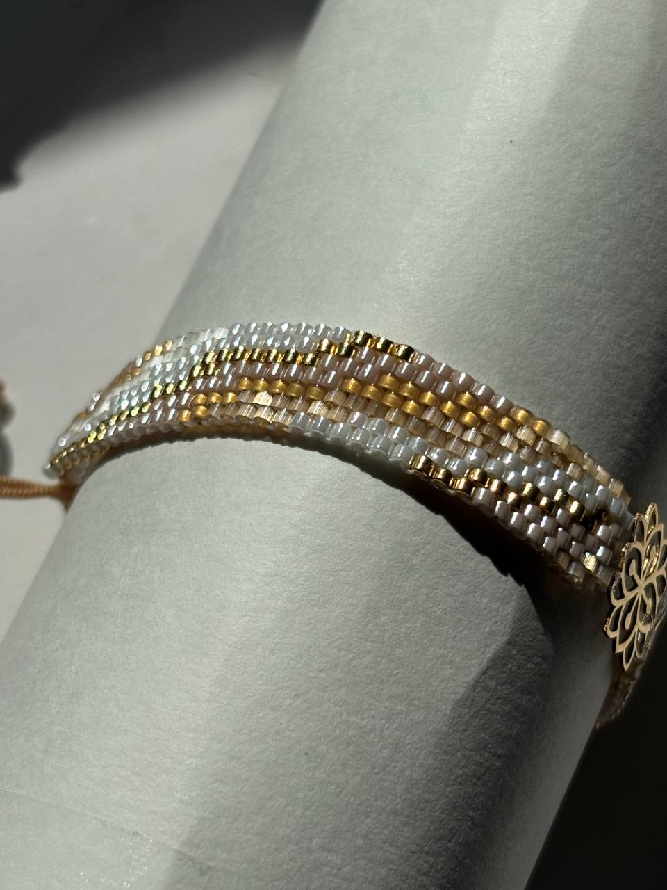 A close-up of a beaded bracelet with white, gold, and silver beads, displayed on a light gray cylindrical stand.