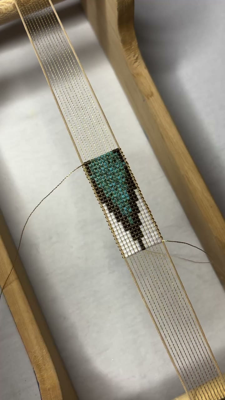 A handmade beaded bracelet with a geometric pattern in turquoise, brown, and white, finished with a gold-toned adjustable cord closure, resting on draped cream fabric.
